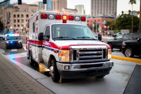 An ambulance with flashing lights is parked on a city street, while police vehicles are visible in the background.