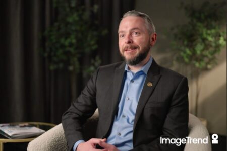 A man in a suit sits in a chair, smiling, with a blurred background of plants. A magazine rests on a side table. The ImageTrend logo appears in the lower right corner.