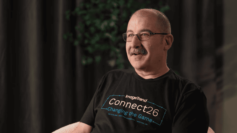 A man with glasses and a mustache sits indoors, wearing a black "ImageTrend Connect26" T-shirt. A plant and dark curtains are visible in the background.