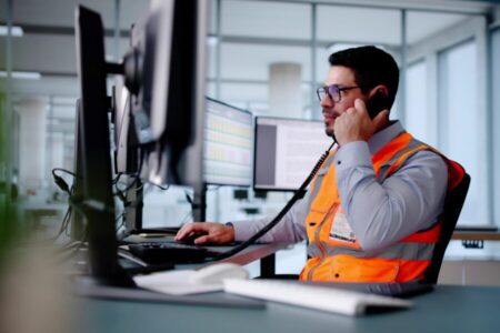 A man wearing an orange safety vest and glasses sits at a desk with multiple monitors, talking on a landline phone in a modern office setting.