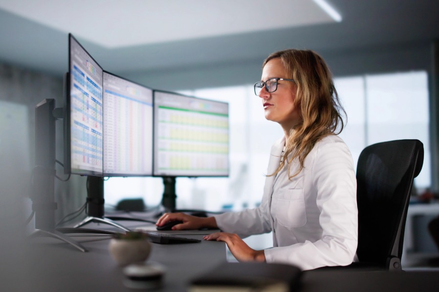 A woman wearing glasses sits at a desk working on a computer with multiple large monitors displaying data in an office setting.