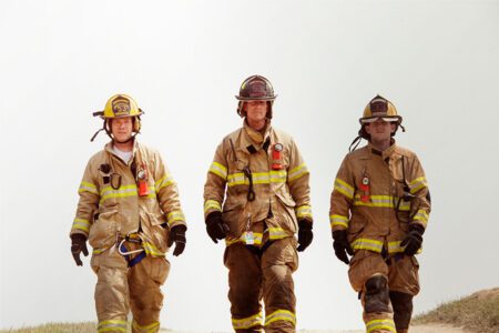 Three firefighters in full gear walk side by side outdoors on dirt, wearing helmets and reflective uniforms, with a pale sky background.