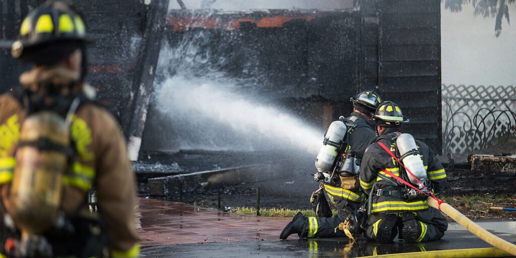 Three firefighters in full gear spray water onto a burning building, while another firefighter stands nearby observing the scene.