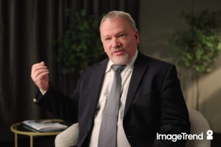 A man in a suit sits in a chair, gesturing with his hand while speaking about EMS documentation. A small table and plants are in the background. The ImageTrend logo is displayed in the lower right corner.