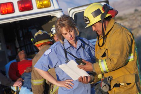 A firefighter and a paramedic consult a clipboard next to an ambulance, while another paramedic tends to an injured person on a stretcher in the background.