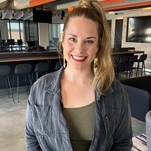 Woman with long blonde hair in a ponytail, wearing a gray jacket and olive shirt, smiling in a modern office with bar stools and large windows in the background.