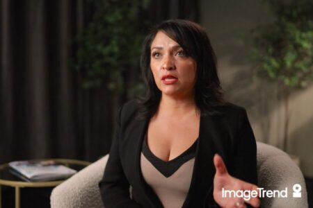 A woman with dark hair sits in a chair, speaking with one hand raised, against a blurred background with plants and a table—capturing the focus often required in EMS documentation. The ImageTrend logo appears in the lower right corner.
