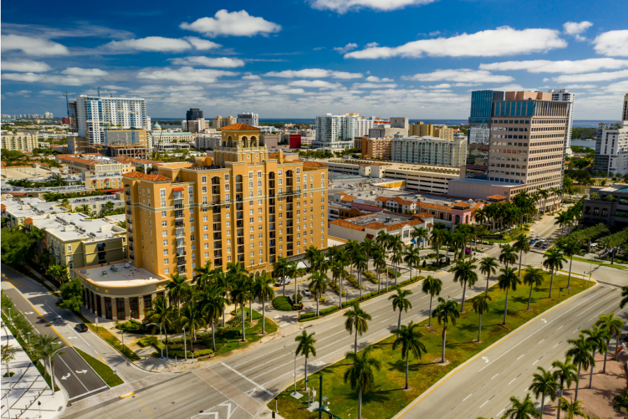 Aerial view of a cityscape with tall buildings, palm trees lining the streets, and a partly cloudy sky overhead.