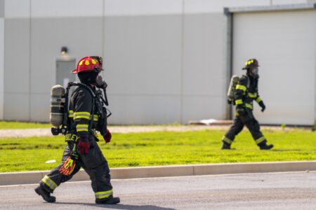 Two firefighters in full gear and helmets walk on a paved road beside a grassy area, with a large industrial building in the background, possibly discussing recent EMS software implementation.