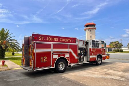 A red St. Johns County fire truck marked “12” is parked outside near a road, with a watchtower and palm trees in the background under a blue sky, as part of the county’s new AI-assisted emergency response system.