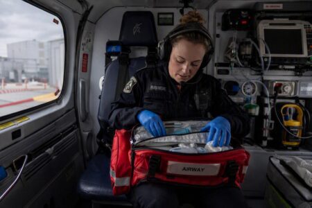 A paramedic wearing gloves sits in a medical helicopter, preparing airway equipment from a red emergency bag.