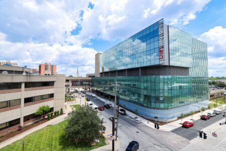 The modern glass facade of the AI-integrated UofL Hospital building viewed from an elevated angle above a street intersection.