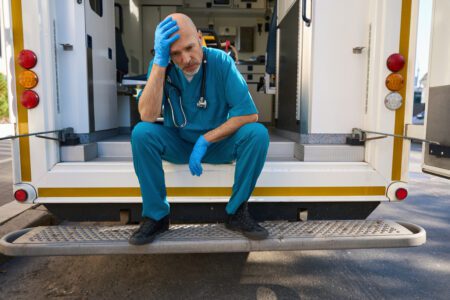 A weary paramedic in scrubs and blue gloves sitting on the back of an ambulance with his head in his hand, an image often seen in EMS World.