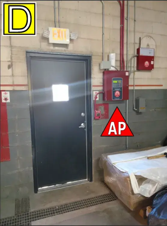 Industrial area with a black exit door, warning symbols, and fire safety equipment on a cinder block wall.