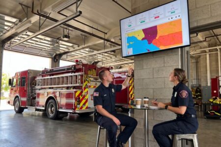 Two firefighters in a fire station look at a wall-mounted digital map display showing regional statistics and locations.