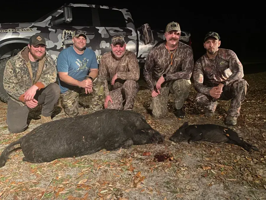 Six men in camouflage kneel outside at night beside two harvested wild hogs, with a camo-patterned truck in the background.