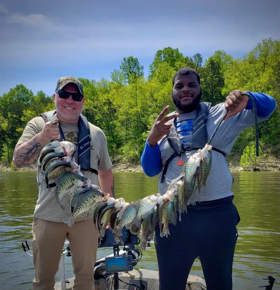 Two men on a boat hold up a long line of caught fish, smiling at the camera. Trees and water are visible in the background.
