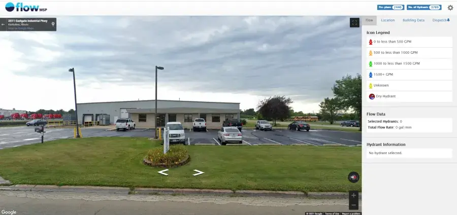 A single-story industrial building with several parked cars in front, surrounded by a parking lot and grass, under a cloudy sky.