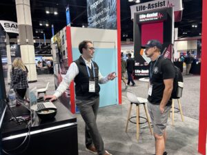 A man in a black vest and glasses gestures while talking to a man in a black t-shirt and baseball cap at a NERIS trade show booth.