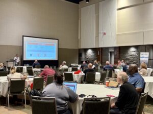 A NERIS speaker presents in front of a conference room audience seated at round tables.