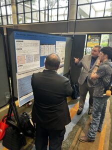 Two men in business attire and a younger man with a backpack discuss a NERIS medical research poster.