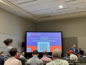 A speaker stands before an audience in a conference room with a large NERIS screen displaying instructions and a QR code for claiming continuing education credits.