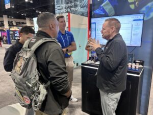 A man in a black pullover gestures while speaking to two men at a NERIS trade show booth featuring a large computer screen.