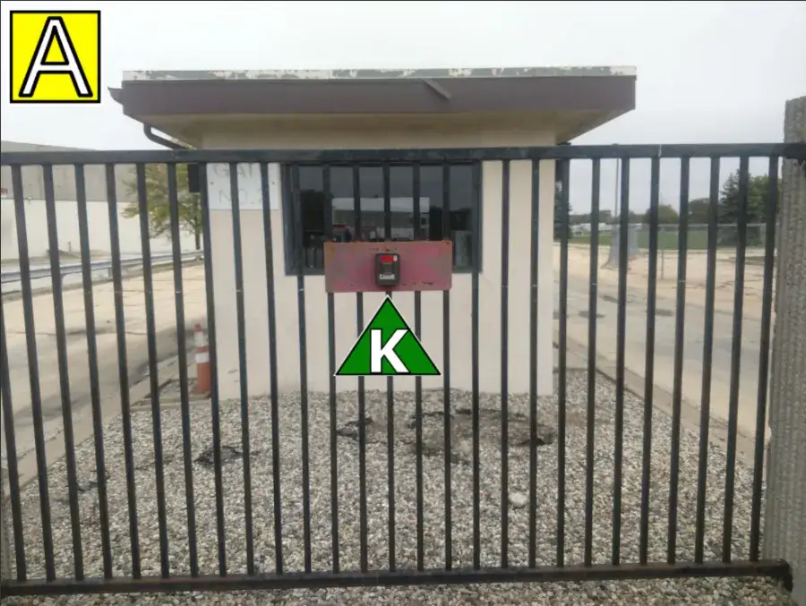 A black metal gate with a keypad access box in front of a small white guard booth. There is a green triangle with a “K” and a yellow square with an “A” in the image.
