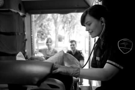 An EMT uses a stethoscope to examine a sickle cell patient on a stretcher inside an ambulance, while two people stand in the background.