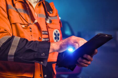 A paramedic in an orange high-visibility uniform uses a tablet for connecting fire and EMS data while holding a small flashlight.